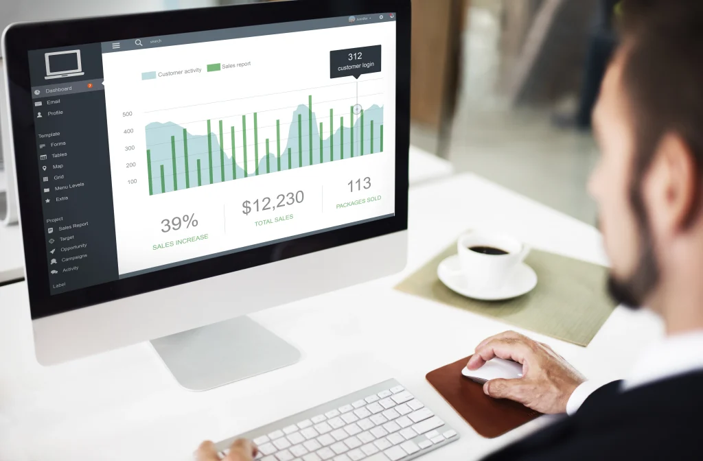 A man in a suit using a computer displaying a business analytics dashboard with green bar charts, customer activity area graphs, and sales data metrics.